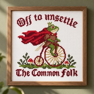May include: A framed cross-stitch embroidery featuring a green frog wearing a crown and a red cape, riding a penny-farthing bicycle. The text reads "Off to unsettle The Common folk" in dark red, with small green plants and red mushrooms below.