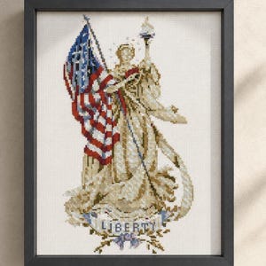 Può includere: Opera d'arte a punto croce incorniciata raffigurante la Statua della Libertà con una bandiera americana e una torcia. L'opera utilizza fili beige, rossi, bianchi e blu, con la parola "LIBERTY" in basso. La cornice è nera.