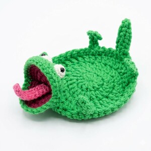 May include: A handmade green crochet creature with a wide open mouth and a pink tongue. The creature has white eyes and is lying on its back. The crochet work is detailed and the overall design is whimsical.