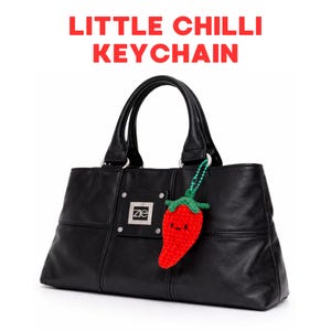 May include: Black leather handbag with a red crocheted chilli pepper keyring. The keyring has a green stem and a smiling face. The text "LITTLE CHILLI KEYCHAIN" is displayed above the bag.