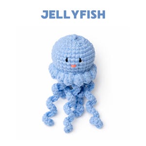 Pode incluir: Um brinquedo de água-viva de crochê azul claro com uma cabeça redonda, bordas com babados e longos tentáculos encaracolados. A água-viva tem pequenos olhos pretos e uma boca cor de pêssego. A palavra "JELLYFISH" está impressa em azul acima do brinquedo.