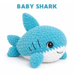 May include: A crocheted baby shark plush toy in shades of blue and white. The toy has a round body, fins, and a smiling face with black eyes. The text "BABY SHARK" is above the toy.