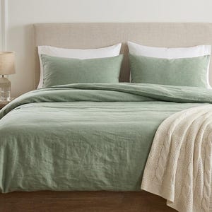 Op de afbeelding: Een bed met saliegroene beddengoed, inclusief een dekbedovertrek en kussenslopen. Een crèmekleurige kabelgebreide sprei is over het voeteneinde van het bed gedrapeerd. Een klein houten nachtkastje met een lamp en plant staat naast het bed.