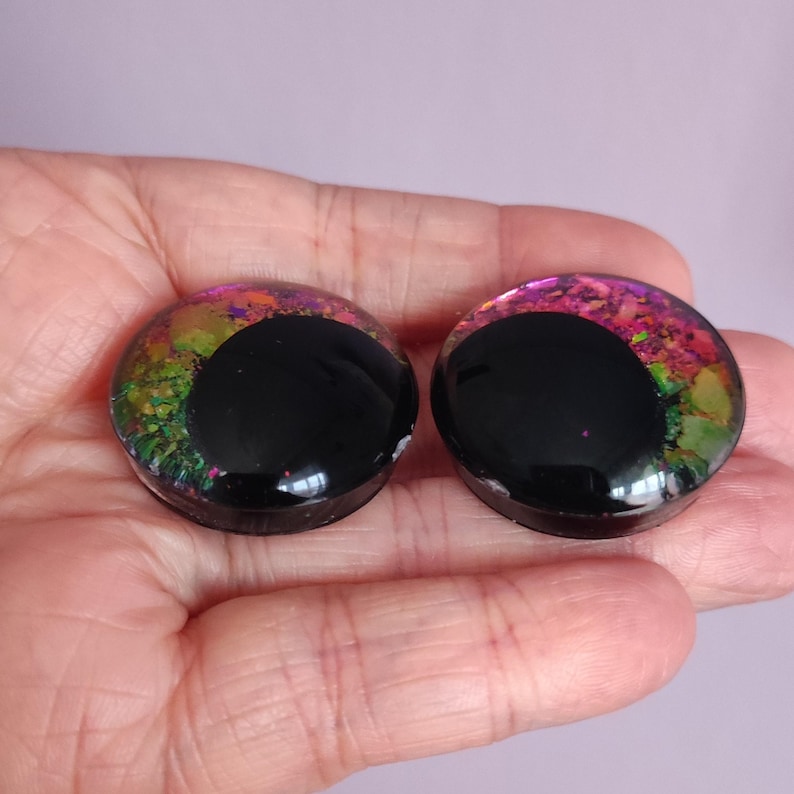 Puede incluir: Un par de expansores de oreja circulares con un acabado negro liso y brillante en la parte frontal, acentuado por un patr&oacute;n de escamas iridiscentes multicolores en tonos rosa, verde y amarillo visible en los bordes exteriores de los discos.