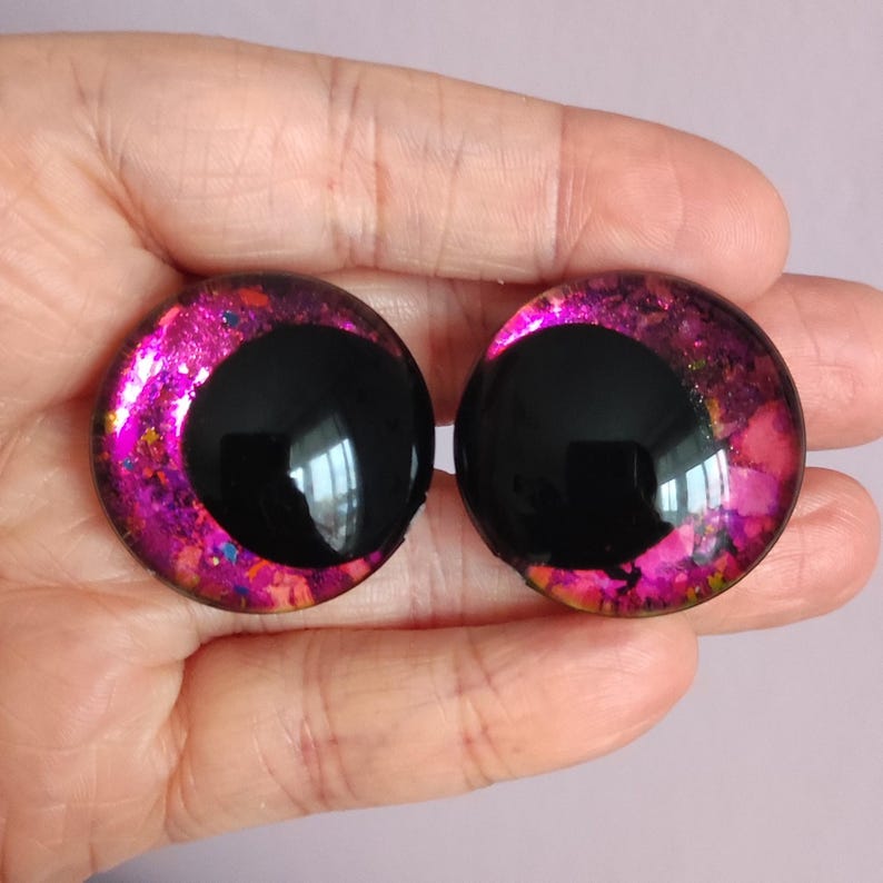 Puede incluir: Dos cabujones circulares con una pupila negra grande y s&oacute;lida sobre un iris vibrante de m&uacute;ltiples tonos. El iris muestra una mezcla de magenta brillante, morado intenso y motas doradas brillantes, creando una apariencia reflectante similar al vidrio.
