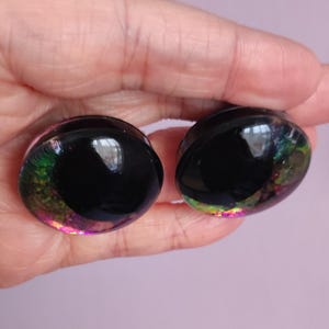 Puede incluir: Un par de expansores de oreja circulares con un centro negro s&oacute;lido rodeado por un anillo exterior transl&uacute;cido lleno de escamas de papel iridiscente multicolor que brillan en tonos verde, morado y dorado sobre una base de resina transparente.