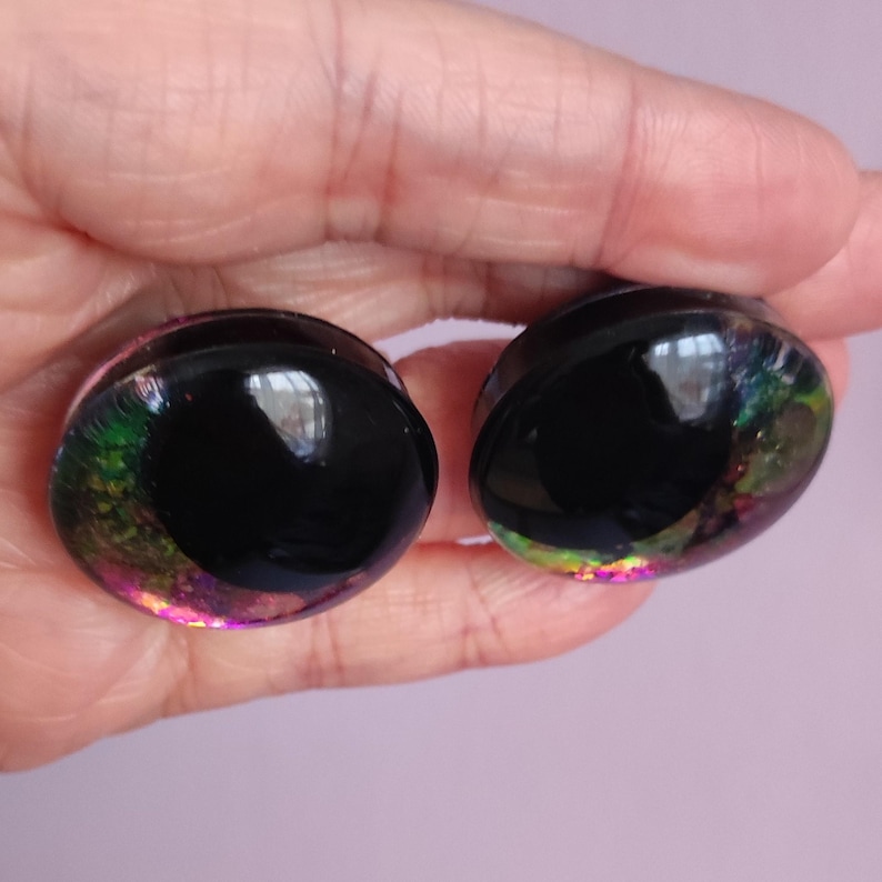 Puede incluir: Un par de expansores de oreja circulares con un centro negro s&oacute;lido rodeado por un anillo exterior transl&uacute;cido lleno de escamas de papel iridiscente multicolor que brillan en tonos verde, morado y dorado sobre una base de resina transparente.
