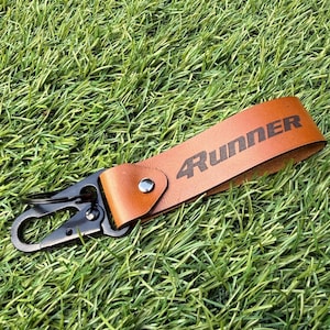 以下が含まれることがあります： 黒で4Runnerの文字が刻印されたタンカラーのレザーキーホルダー。レザーストラップは折りたたまれ、銀色の金属製リベットで固定されています。黒い金属製のカラビナクリップと円形の二重リングがレザーのループに取り付けられています。