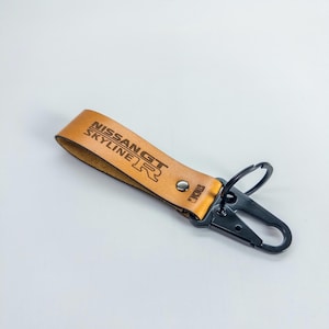 May include: Tan leather keychain with the text "NISSAN GT SKYLINE R" in black. The keychain has a black metal clasp and a silver rivet. The background is a solid white.