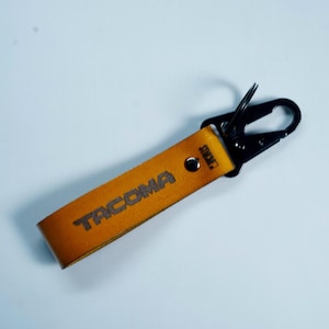 May include: A tan leather keychain strap featuring the word TACOMA embossed in dark lettering. The strap is secured with a silver-toned metal rivet and attached to a black metal carabiner clip with a small circular split ring for keys.