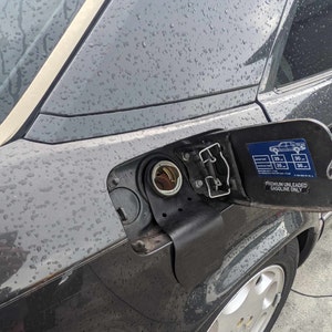 Puede incluir: Primer plano de la tapa del tanque de gasolina de un coche gris con una tapa de combustible negra. La tapa tiene una pegatina que dice "Premium sin plomo, gasolina solamente".