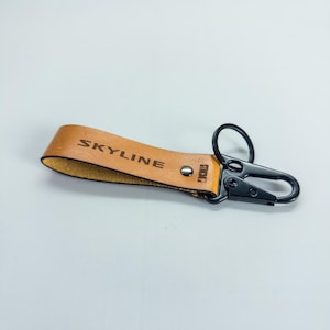 May include: A tan leather keychain with the word "SKYLINE" embossed on the strap. The keychain has a black metal clasp and a small metal rivet. The word "STICKO" is also visible on the strap.