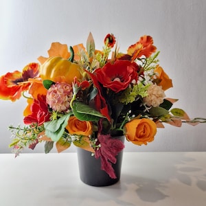 May include: A seasonal floral display in a black pot, featuring orange and red poppies, roses, and a small yellow pumpkin. The arrangement includes green foliage and accents, creating an autumnal theme. The pot is approximately 15 cm in height.