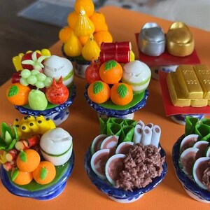 Pode incluir: Miniaturas de alimentos, incluindo arranjos de frutas, barras de ouro e objetos em forma de moeda. Os arranjos de frutas apresentam laranjas, uvas e outras frutas em pratos com estampas azuis e brancas.