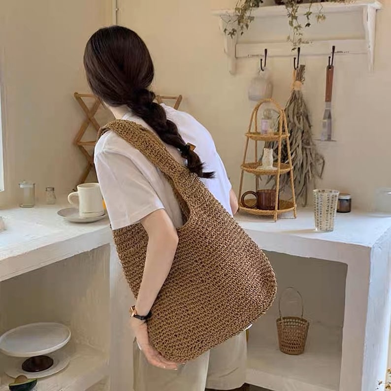 May include: A tan-coloured woven shoulder bag. The bag has a large, open design with a wide strap. The texture of the bag appears to be made of a straw-like material, suitable for carrying items.