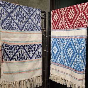 Puede incluir: Dos textiles tejidos con patrones geométricos, uno azul y blanco, el otro rojo y blanco, colgados de un soporte de metal negro. Ambos presentan un borde crema y flecos, mostrando sus intrincados diseños.