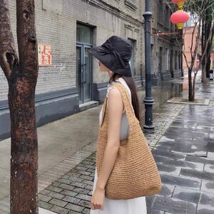 May include: A woman wearing a black sun hat with a bow and carrying a woven straw tote bag. The bag is light brown and has a shoulder strap. The woman is wearing a white dress.