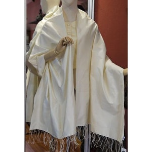 May include: A cream-colored shawl with a fringed edge is draped over a mannequin. The shawl is made of a smooth, flowing fabric. The mannequin is wearing a matching cream-colored top.