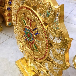 Könnte beinhalten: Ein goldenes, verziertes Dharma-Rad mit komplizierten Details und bunten Edelsteinen. Das Rad ist auf einem Holzsockel montiert und von einem Lotusblumenmuster umgeben.