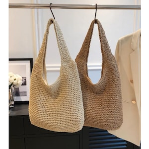 May include: Two woven shoulder bags in beige and brown hang from a metal rod. The bags have a large, open design with long straps. The bags are made of a textured, woven material, and are suitable for carrying items.