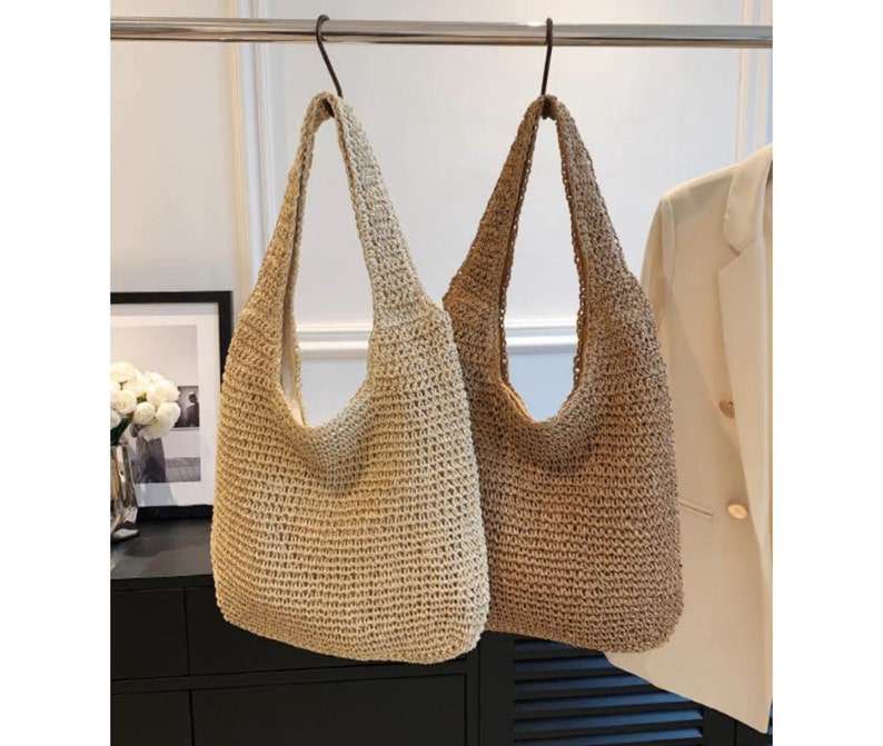May include: Two woven shoulder bags in beige and brown hang from a metal rod. The bags have a large, open design with long straps. The bags are made of a textured, woven material, and are suitable for carrying items.