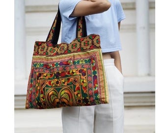 May include: A colorful tote bag with a detailed, patterned design in shades of red, green, yellow, and purple. The bag has black straps and a rectangular shape, suitable for carrying various items. The bag's design features floral and geometric elements.