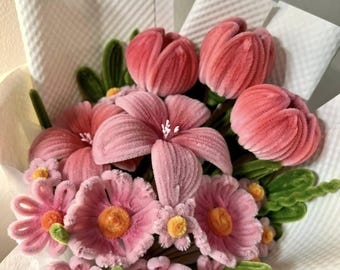 May include: A bouquet of handmade pink flowers, including tulips, lilies, and daisies, wrapped in white paper. The flowers have a soft, velvety texture and are accented with green stems and leaves. The bouquet is a charming, handcrafted floral arrangement.