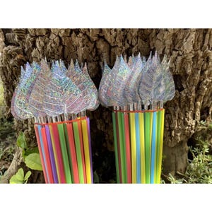 Könnte beinhalten: Zwei Bündel dekorativer Strohhalme mit blattförmigen Glitzertoppern. Die Strohhalme sind mehrfarbig, darunter rot, grün, gelb und blau. Die Blattoberteile sind transparent mit schillerndem Glitzer, wodurch ein schimmernder Effekt entsteht. Der Hintergrund zeigt einen Baumstamm und Grün.
