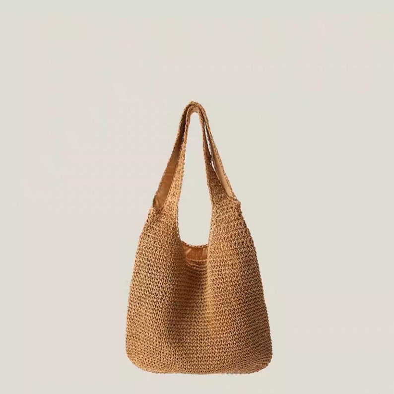 May include: A light brown woven straw tote bag with long handles. The bag has a spacious interior. The handles are reinforced with a darker brown material. The bag is designed for carrying items.