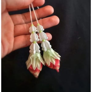 Pode incluir: Duas guirlandas florais, cada uma com flores brancas em forma de sino, detalhes verdes e botões com pontas vermelhas. As guirlandas são enfiadas em cordões brancos, criando um design ornamental delicado.