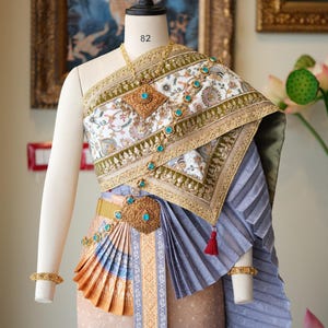 May include: A traditional Thai dress displayed on a mannequin. The dress features a white and gold top with turquoise accents, a pleated gray skirt, and a gold belt. The dress is adorned with intricate embroidery and jewelry.