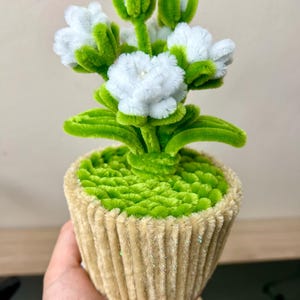 May include: A handcrafted floral display in a cupcake-shaped pot. The flowers are white with green stems and leaves. The pot is tan and green, with a textured, ribbed design. A unique decorative item.