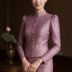May include: A mauve-colored traditional Thai outfit, featuring a high-collared jacket with gold buttons and a matching skirt. The fabric has a subtle geometric pattern. The outfit is worn by a person with dark hair and a serene expression.