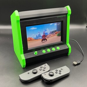 Puede incluir: Una consola de juegos de estilo arcade verde y negro con una pantalla que muestra un juego de carreras de dibujos animados. La consola tiene seis botones y una palanca de mando. Dos mandos grises de Nintendo Switch están en primer plano.