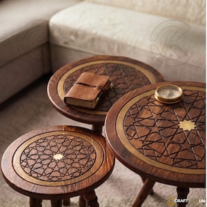May include: A set of three round wooden side tables featuring intricate geometric star patterns carved into the surface, accented with brass circular inlays and a central star. A brown leather-bound notebook and a brass compass rest on top.