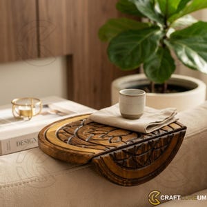 May include: A wooden sofa arm tray with carved floral designs. The tray is brown and has a built-in handle. A small beige cup and a folded beige cloth are on the tray. The tray is on a beige sofa.