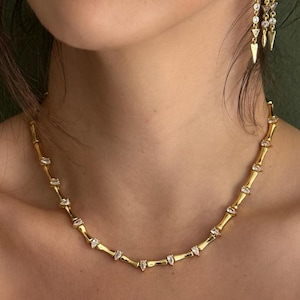 May include: A gold necklace with small, rectangular accents and a matching pair of gold earrings with dangling, pointed charms. The jewelry is worn with a white button-down shirt.