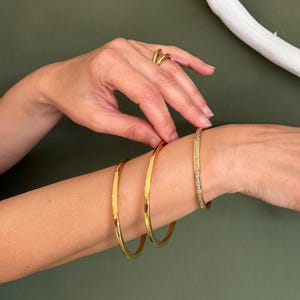 Puede incluir: Un conjunto de tres pulseras doradas, que incluye dos brazaletes lisos y pulidos y uno estrecho con textura, junto con tres anillos dorados en los dedos. Las piezas de joyería presentan un acabado metálico y un diseño circular sencillo.