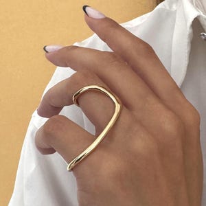 Könnte beinhalten: Ein goldfarbener Ring mit einem einzigartigen, geschwungenen Design. Der Ring wird an einem Finger getragen, und das Design verläuft über die Oberseite des Fingers. Ein weißes Hemd und eine goldene Halskette sind ebenfalls sichtbar.
