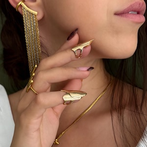 Op de afbeelding: Goudkleurige sieradenset met een oormanchet met bungelende kettingen, twee vingerringen en een ketting met een hanger. De ringen hebben een uniek, hoekig ontwerp. De sieraden worden gedragen met een wit overhemd.
