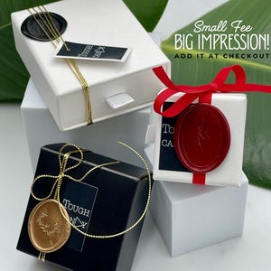 three gift boxes with various designs and colors, including a white box with a red ribbon, a black box with a gold ribbon, and a white box with a red ribbon. The boxes are placed on a white surface, and there is a green leaf in the background.