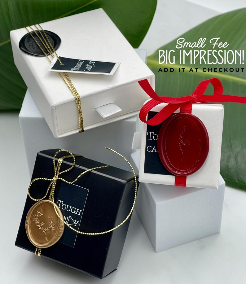 three gift boxes with various designs and colors, including a white box with a red ribbon, a black box with a gold ribbon, and a white box with a red ribbon. The boxes are placed on a white surface, and there is a green leaf in the background.