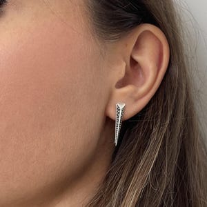 Peut inclure: Une boucle d'oreille géométrique argentée avec un sommet triangulaire et une longue forme pointue. La boucle d'oreille présente un motif de petits points sombres le long de ses bords. La boucle d'oreille est portée à l'oreille.