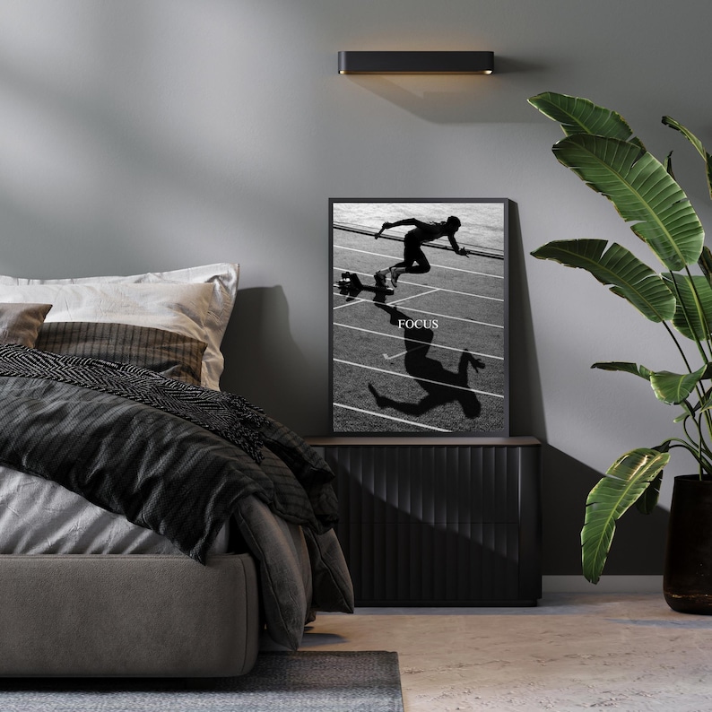 May include: Black and white framed print featuring a runner in motion on a track, with the word "FOCUS" in white text. The print is displayed on a dark wood cabinet, with a large green plant to the right.