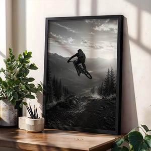 May include: A black and white framed wall art print depicting a mountain biker mid-air during a jump. The scene features a silhouette of the rider and bike against a backdrop of layered mountain ranges, pine trees, and a cloudy sky at sunset.