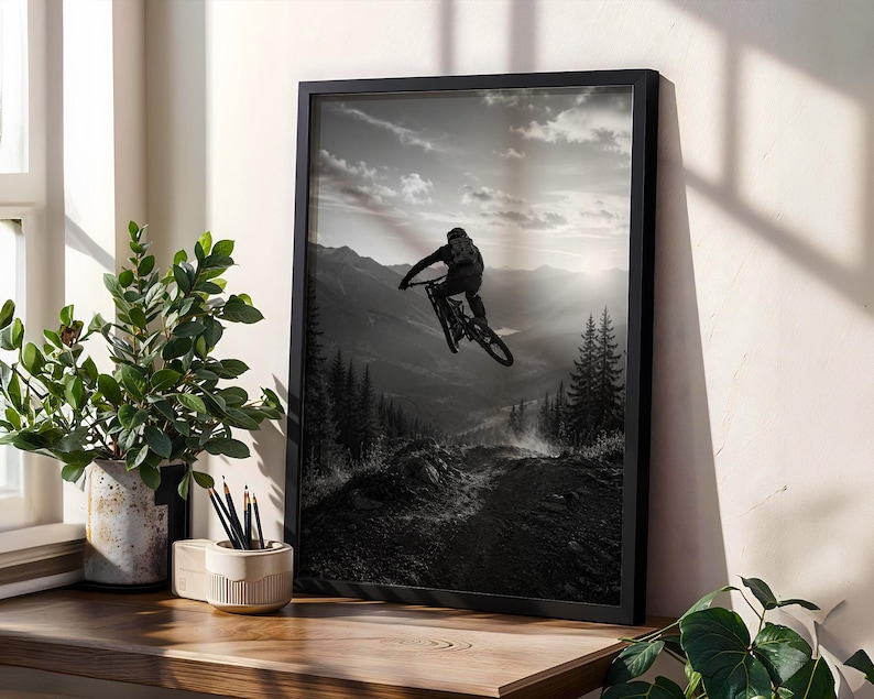 May include: A black and white framed wall art print depicting a mountain biker mid-air during a jump. The scene features a silhouette of the rider and bike against a backdrop of layered mountain ranges, pine trees, and a cloudy sky at sunset.