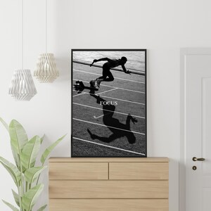 May include: Black and white framed print featuring a sprinter in motion on a track. The word "FOCUS" is printed in white at the bottom of the image. The print is displayed on a wooden dresser.
