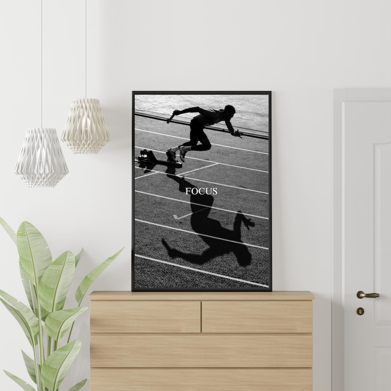 May include: Black and white framed print featuring a sprinter in motion on a track. The word "FOCUS" is printed in white at the bottom of the image. The print is displayed on a wooden dresser.