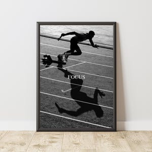 May include: Black and white framed print of a sprinter in motion on a track. The word "FOCUS" is printed in white below the runner. The runner's shadow is cast on the track.