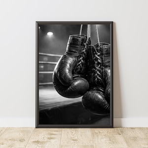 Può includere: Stampa incorniciata in bianco e nero con un paio di guantoni da boxe vintage appesi a delle corde. I guantoni sono usurati e presentano lacci visibili. Lo sfondo mostra un ring di boxe e un riflettore.